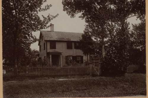 A vintage black-and-white photo of a two-story house surrounded by trees and a picket fence.