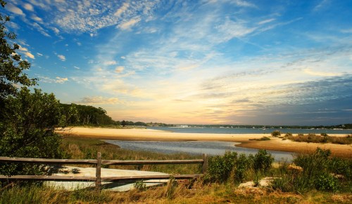 A serene beach scene at sunset, featuring a calm waterway, sandy shore, and lush greenery under a colorful sky.