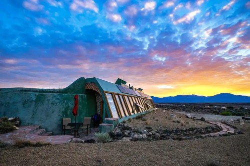 A unique green building with large windows, set against a colorful sunset and mountains in the background.