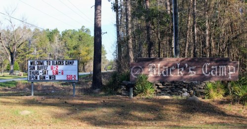 Sign for Black's Camp with hours and buffet details, surrounded by trees and a gravel road.