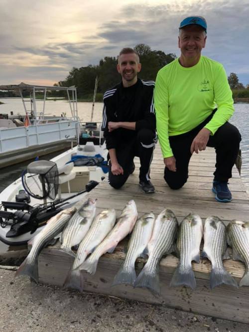 Two men kneel beside a row of freshly caught fish on a dock, with boats and a cloudy sky in the background.