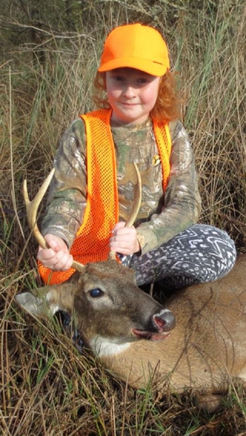A young child in an orange vest and hat sits in grass, holding antlers of a deer they have hunted.
