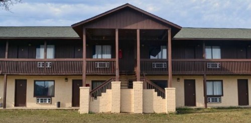 Two-story motel with a wooden balcony, multiple doors, and air conditioning units under a cloudy sky.