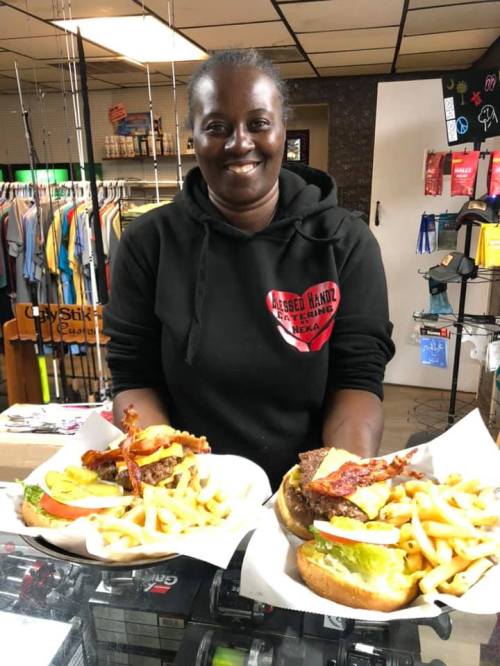 A smiling woman holds two plates of burgers with fries, standing in a casual restaurant setting.