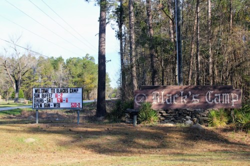 Sign for Black's Camp with operating hours and a welcome message, surrounded by trees and grass.