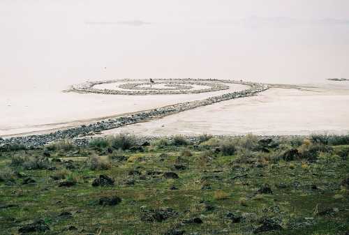 A spiral stone formation on a salt flat, surrounded by sparse vegetation and a hazy sky in the background.