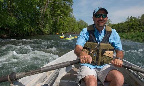 A man in a blue shirt and sunglasses rows a boat on a river, with kayakers visible in the background.