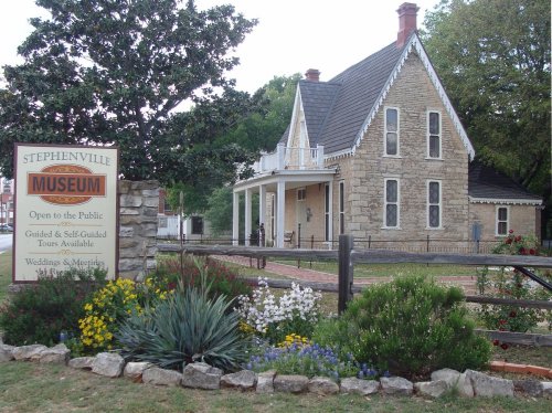 Historic stone building with a sign for Stephenville Museum, surrounded by colorful flowers and greenery.