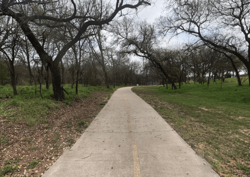 A paved path winds through a wooded area with sparse trees and green grass on either side under a cloudy sky.