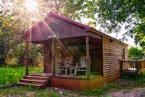 A cozy log cabin with rocking chairs on the porch, surrounded by greenery and sunlight filtering through the trees.