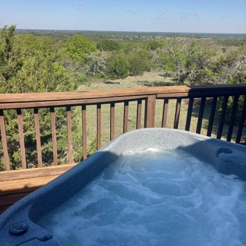 A hot tub on a wooden deck overlooking a scenic landscape of trees and rolling hills under a clear blue sky.