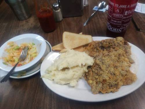 A plate with fried fish, mashed potatoes, toast, and a side salad on a wooden table. A drink is in the background.