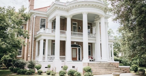 A grand two-story house with white columns, a red roof, and lush greenery surrounding the entrance steps.