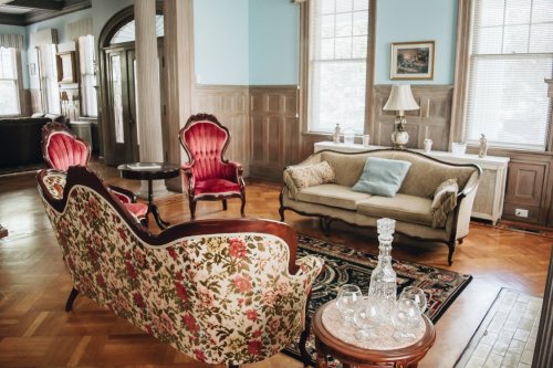 A vintage living room with floral sofas, red velvet chairs, a wooden table, and elegant decor in soft lighting.