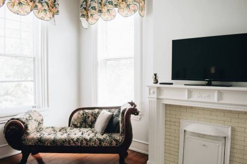 A cozy room featuring a floral chaise lounge, a TV mounted above a white fireplace, and large windows with light curtains.