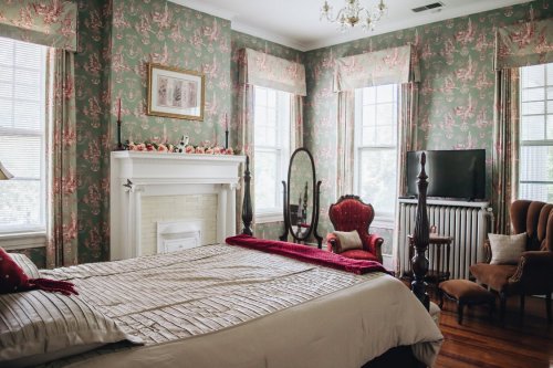 Cozy bedroom with floral wallpaper, a large bed, vintage furniture, and windows letting in natural light.