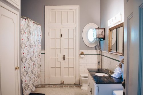 A bright bathroom featuring a shower curtain, toilet, sink, and a round window, with vintage decor and tiled walls.