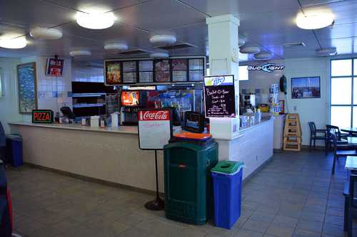 Interior of a casual eatery featuring a counter with menus, seating, and various beverage signs.