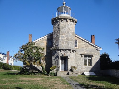 A stone lighthouse with a cylindrical tower attached to a house, set against a clear blue sky.