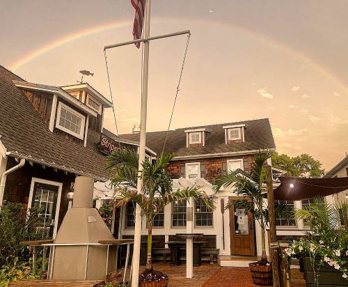A charming restaurant with a flag, palm trees, and a rainbow in the sky, set against a warm sunset backdrop.