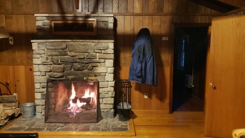 A cozy cabin interior featuring a stone fireplace, wooden walls, and a blue coat hanging by the door.