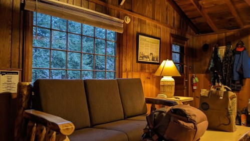 Cozy cabin interior with a wooden sofa, lamp, and large window showing trees outside. A backpack and bags are nearby.