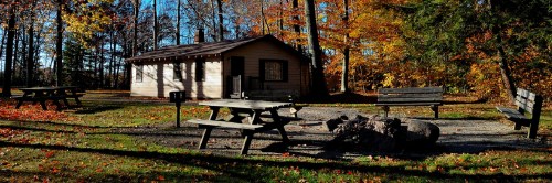 A cozy cabin surrounded by colorful autumn trees, with picnic tables and a fire pit in a serene outdoor setting.