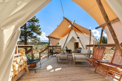 A cozy outdoor seating area in front of a glamping tent, surrounded by trees and open landscape under a blue sky.