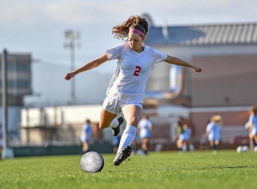A young female soccer player in a white uniform kicks a soccer ball on a grassy field during a game.