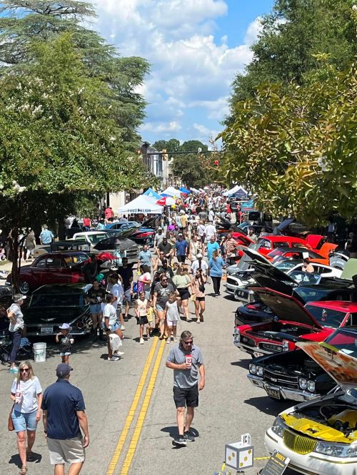 A bustling street filled with people and classic cars during a car show, with tents and trees lining the road.