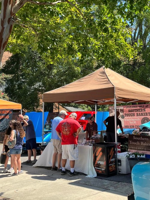 A busy outdoor market scene with people gathered around a tent selling baked goods under a sunny sky.