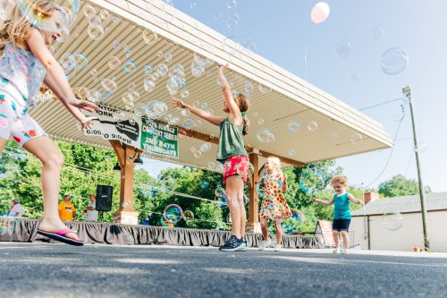 Children playing and dancing under a pavilion, surrounded by floating bubbles on a sunny day.
