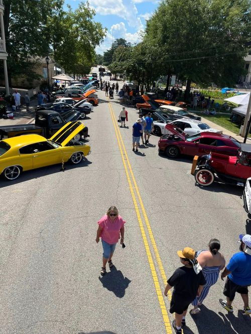 A vibrant car show on a sunny day, featuring classic cars and crowds along a tree-lined street.