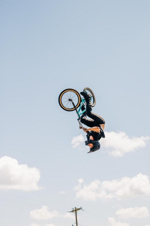 A BMX rider performs a mid-air trick against a clear blue sky with fluffy clouds.