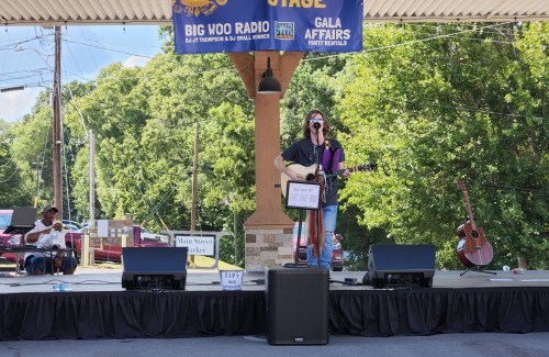 A musician performs on stage with a guitar, while another person sits nearby. Banners hang above in a sunny outdoor setting.