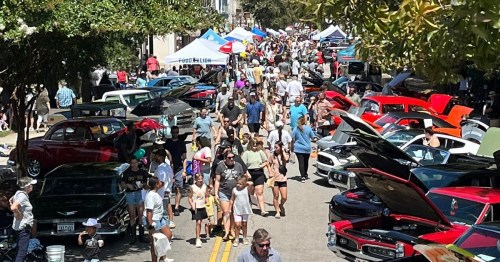A bustling street filled with classic cars and crowds of people enjoying a car show on a sunny day.