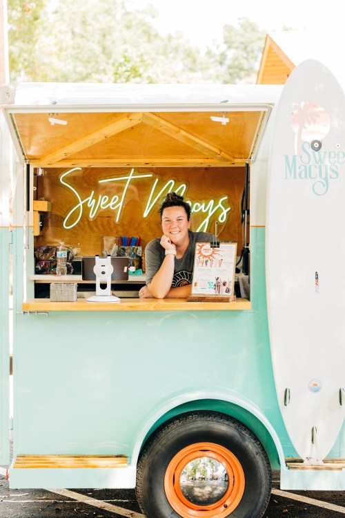 A cheerful vendor leans on the counter of a colorful food truck named "Sweet Mays," with a surfboard decoration.