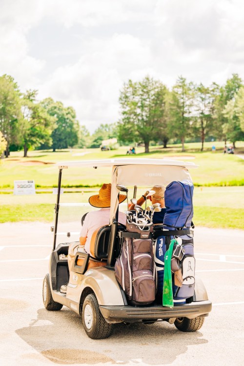 A golf cart loaded with bags and clubs, parked on a sunny golf course with trees in the background.