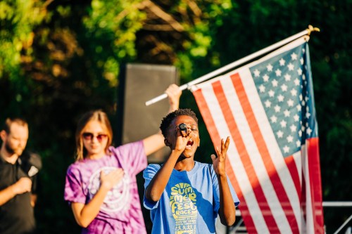 A young boy sings into a microphone while a girl in sunglasses stands behind him, holding an American flag.