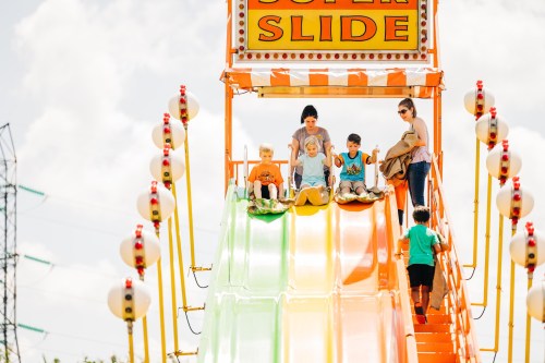Children and adults prepare to slide down a colorful, tall slide at a fair, with bright signage and a sunny sky.