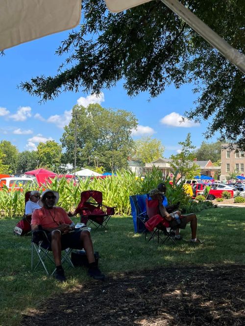 Two people sitting in lawn chairs under a tree, enjoying a sunny day at an outdoor event with tents in the background.