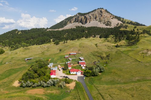 Aerial view of a rural property with red-roofed buildings, surrounded by green fields and a rocky mountain in the background.
