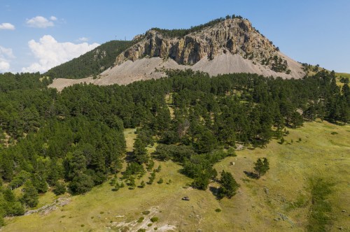 A scenic view of a rocky mountain surrounded by lush green trees and open fields under a clear blue sky.