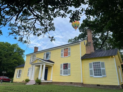 A yellow two-story house with white columns and a hot air balloon floating above it, surrounded by trees and a blue sky.