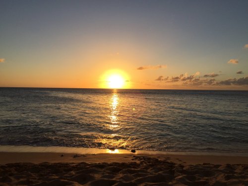 A serene beach at sunset, with the sun reflecting on calm ocean waters and a clear sky.