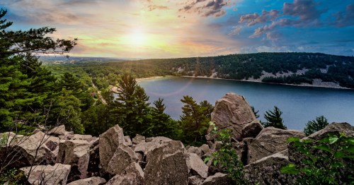 Sunset over a serene lake surrounded by lush green hills under a clear blue sky, with boats dotting the water.