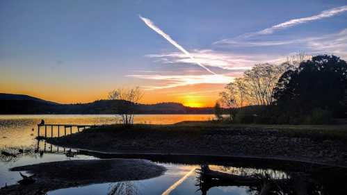 A serene sunset over a lake, with silhouettes of trees and a pier reflecting in the water.