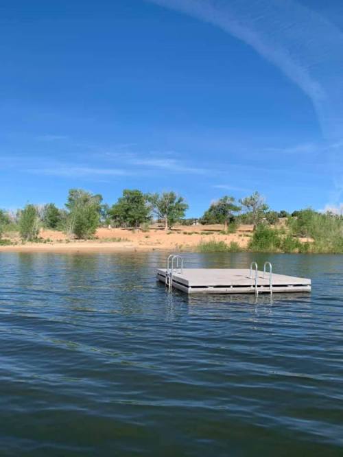 A wooden dock floats on calm water, surrounded by greenery and sandy shores under a clear blue sky.