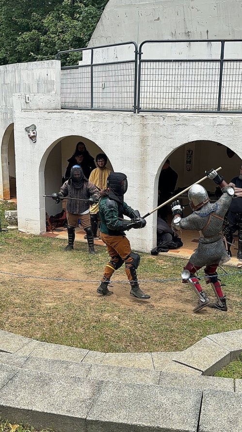 Two armored knights spar with swords in a grassy area, while onlookers watch from a nearby structure.