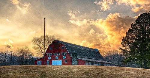A red barn stands on a grassy hill under a dramatic sunset sky with clouds in shades of orange and yellow.
